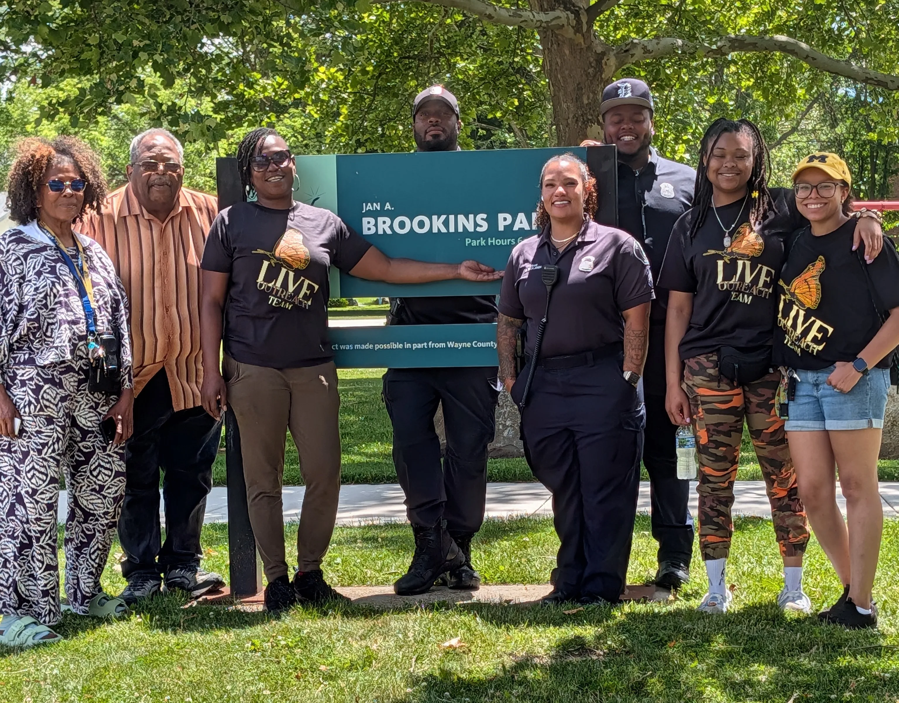 A group of people, several wearing L.I.V.E. Outreach team shirts, standing together and smiling in front of the Jan A. Brookins Park sign in Detroit