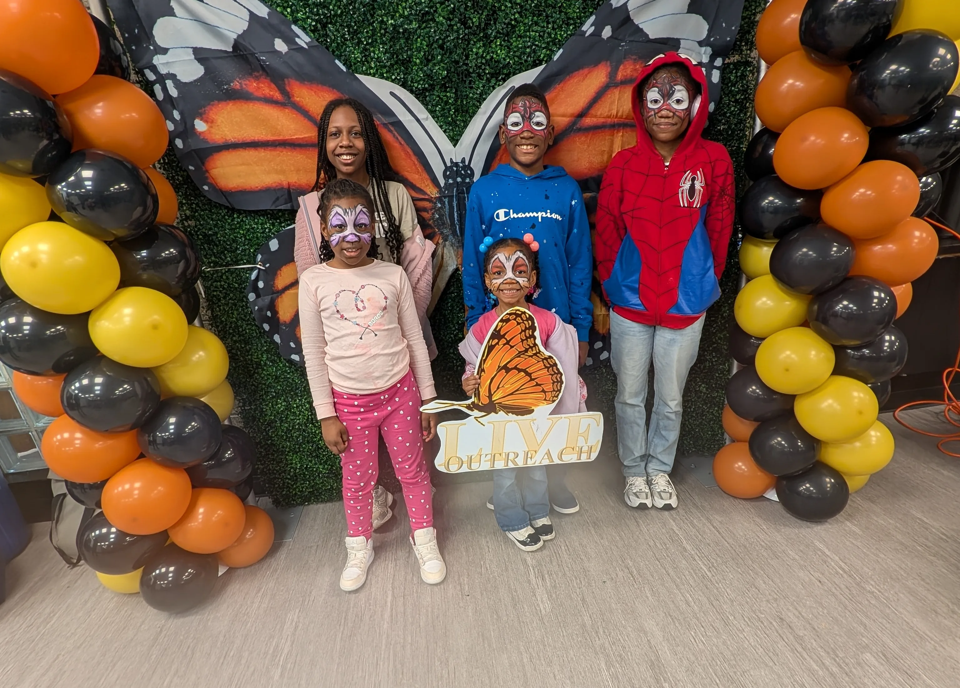Four face-painted kids posing with the L.I.V.E. Outreach butterfly sign in front of a monarch butterfly backdrop and orange and black balloon arches at a community wellness event