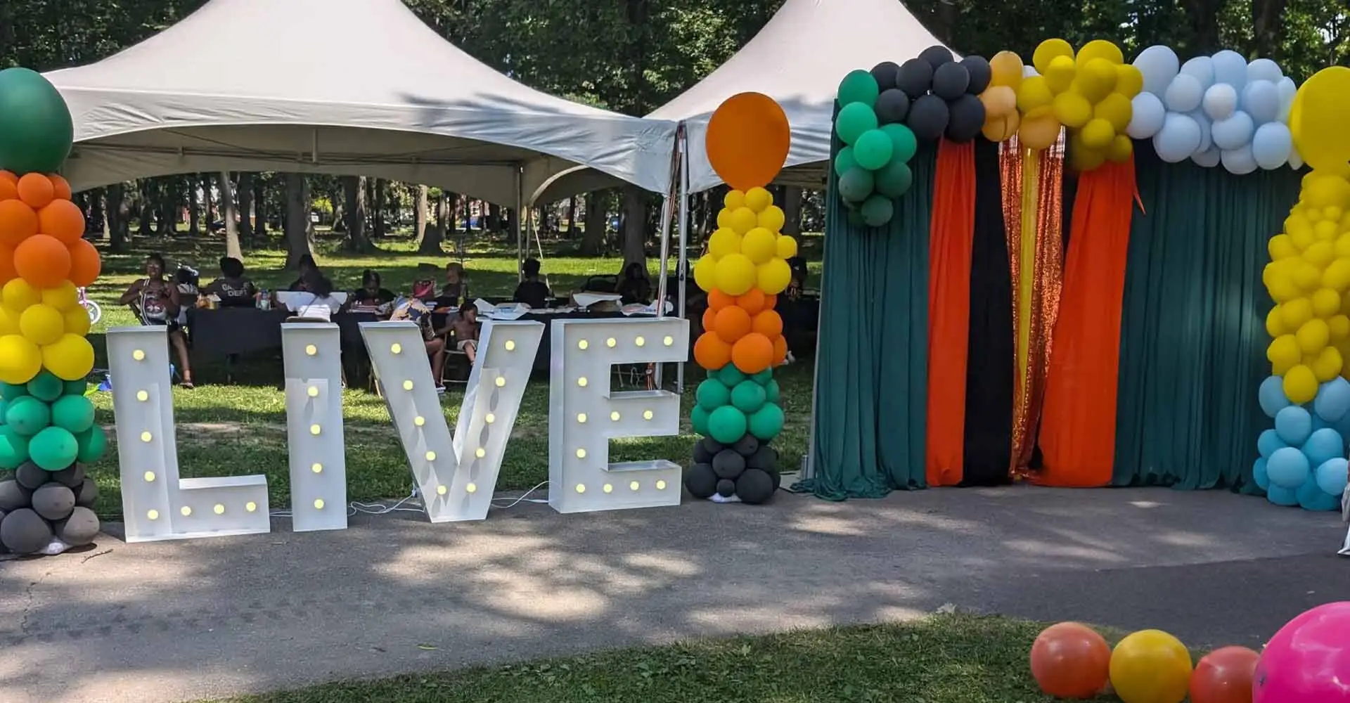 Lit-up LIVE marquee letters with a colorful balloon arch and draped backdrop at an outdoor L.I.V.E. Outreach community event