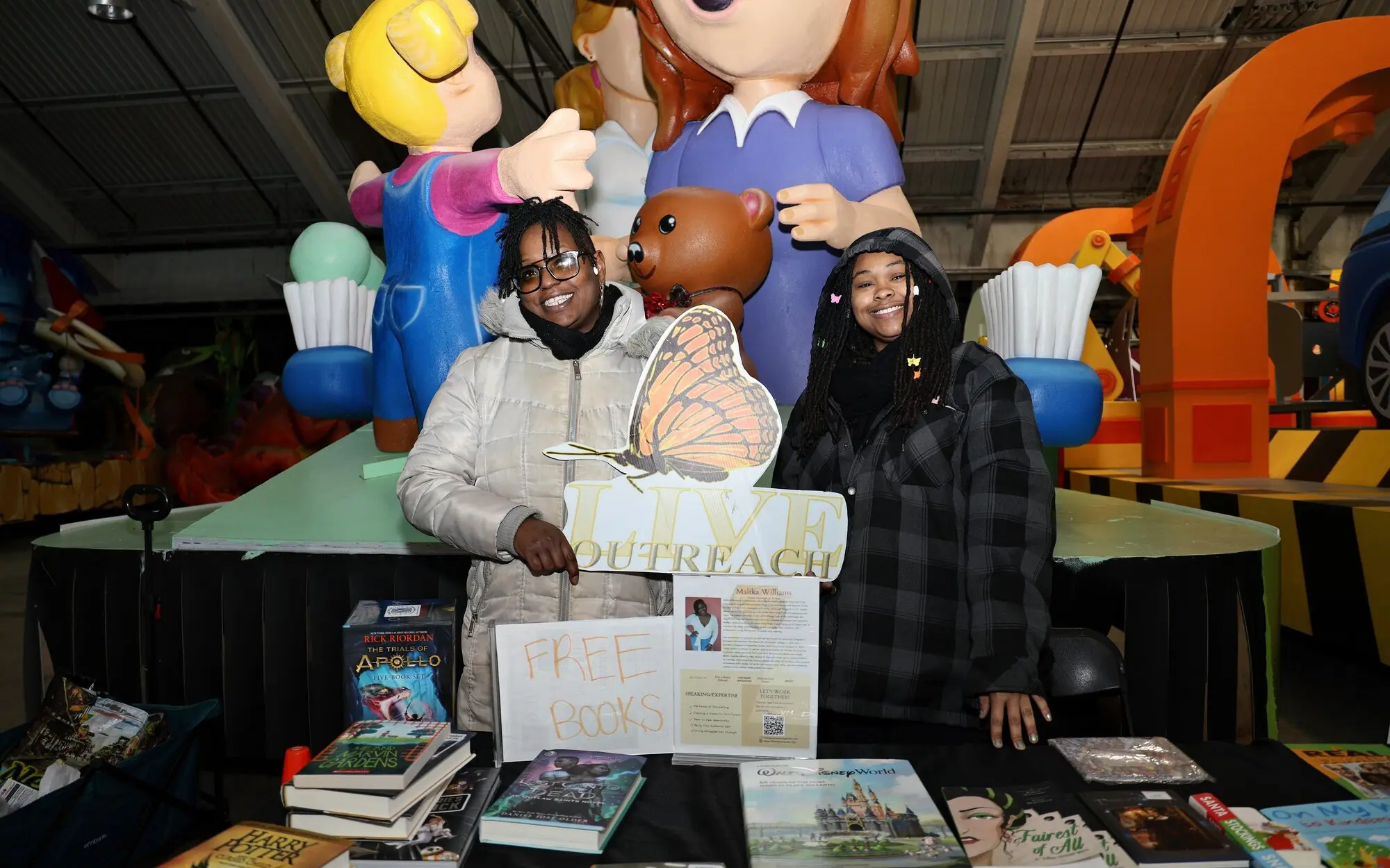 Two L.I.V.E. Outreach volunteers at an indoor book giveaway table with a Free Books sign and the L.I.V.E. Outreach butterfly sign