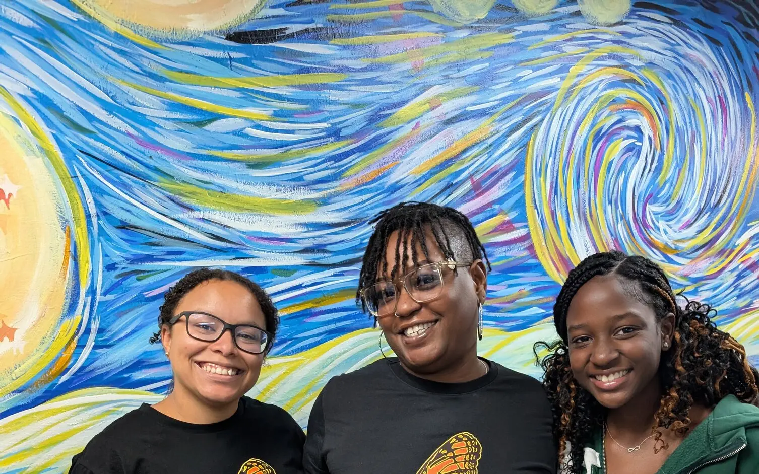 Three L.I.V.E. Outreach team members smiling in branded shirts in front of a colorful mural