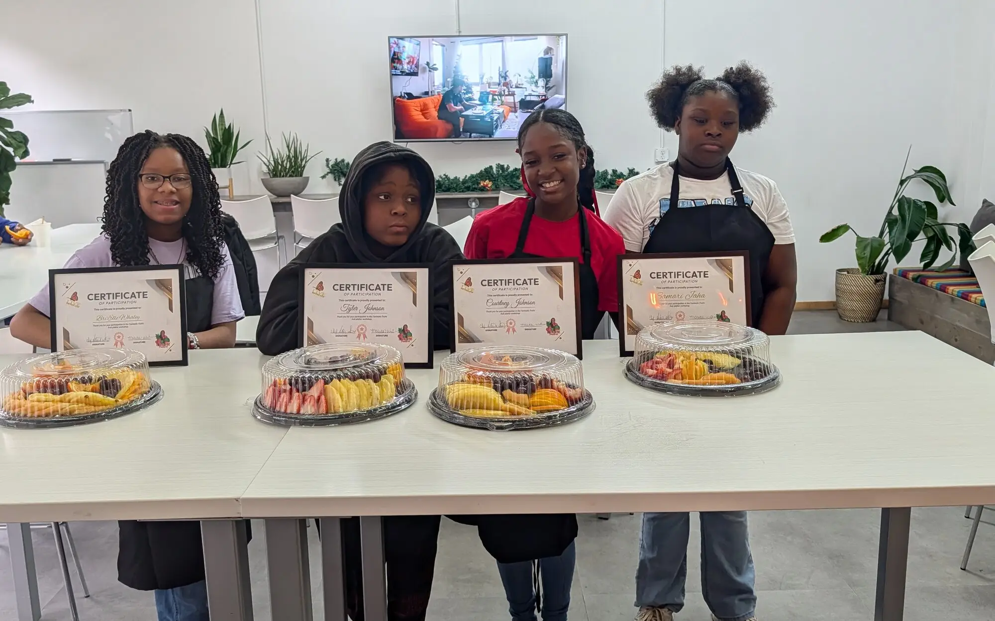 Four youth standing behind a table displaying certificates of participation and fruit platters from a L.I.V.E. Outreach cooking workshop